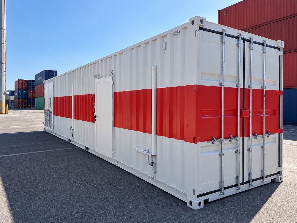 Container industriale modificato bianco e rosso, con porta di accesso laterale e impianti tecnici, in un terminal portuale.