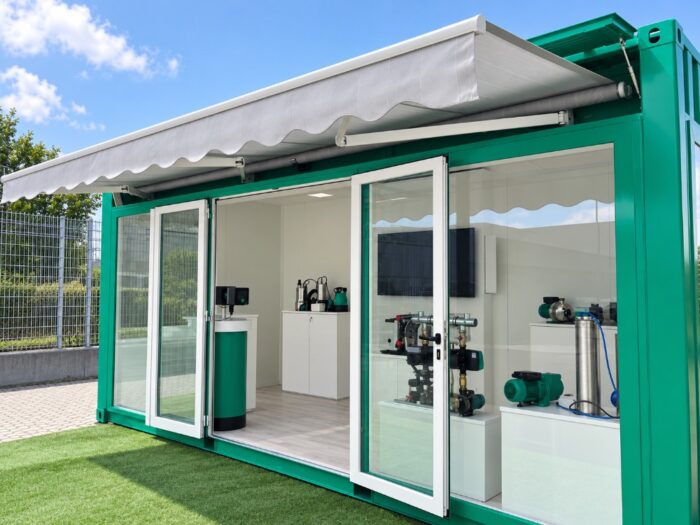 Container trasformato in showroom espositivo, con ampie vetrate, tendalino esterno e allestimento interno per la presentazione di apparecchiature industriali.